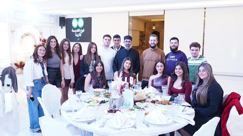 العشاء السنوي لفوج القديسة كاترينا - Scout National Orthodox A large group of young adults is gathered for a formal meal around a circular table in a brightly lit, modern banquet hall. They are smiling at the camera, with some standing behind the table and others seated.